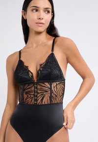Bodysuit negro con detalles de encaje, que cuenta con un corsé de encaje transparente con patrones de hojas y una parte inferior negra sólida. Tirantes ajustables y cierre frontal.