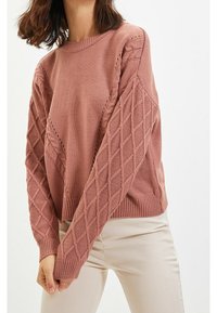 Rosenfarbener gestrickter Pullover mit diamantförmigem Muster an Ärmeln und Seiten, Rundhalsausschnitt und geripptem Saum. Getragen über hellen Hosen.