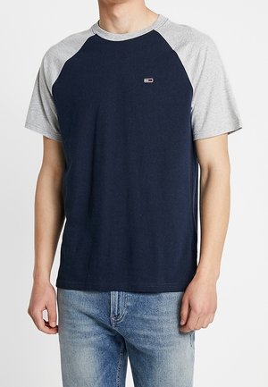 Mand iført en navyblå og lysegrå raglanærmet T-shirt kombineret med lyseblå jeans, stående mod en ensfarvet baggrund.