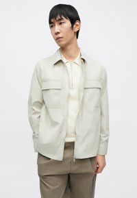 Világos bézs cipzáras dzseki két első zsebbel, vajszínű pólóinggel és világosbarna nadrággal párosítva, sima textúrával.