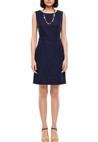 Femme portant une robe bleu marine sans manches longueur genoux, un collier de perles multicolores et des sandales à talons hauts jaunes à brides.