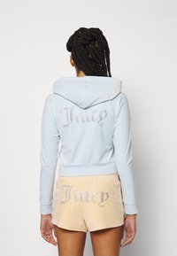 En person med flätat hår bär en ljusblå kort hoodie och persikofärgade shorts, båda med ordet "Juicy" i rhinstenar.