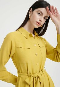 Vestito giallo senape con bottoni e colletto, tasche frontali e dettaglio in vita legato. Maniche lunghe con finitura in tessuto texturizzato.
