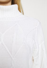 Bílý svetr z pleteniny s vysokým ribovým límcem, který má texturované vzory a otevřené tkaní, předvádějící jemnou, lehkou látku.