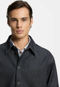 Giovane uomo con capelli scuri indossa una giacca di jeans scura sopra una camicia bianca, fronteggiante con espressione neutra su sfondo chiaro e semplice.