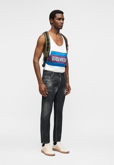Άνδρας φορώντας λευκό αμάνικο τοπ με το λογότυπο «DSQUARED2», σκούρα ξεθωριασμένα τζιν, μπεζ αθλητικά παπούτσια και ελιάς πράσινο σακίδιο, στέκεται σε μονόχρωμο φόντο.