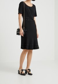 Abito nero con maniche corte, caratterizzato da occhielli in metallo lungo la vita e l'orlo. Abbinato a scarpe nere con tacco a blocco e una borsa piccola.