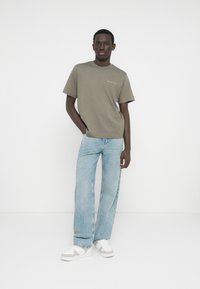 Χακί βαμβακερό t-shirt με κοντά μανίκια και λευκό κείμενο στο στήθος, συνδυασμένο με ανοιχτό μπλε τζιν παντελόνι με φαρδιά πόδια και λευκά αθλητικά παπούτσια.