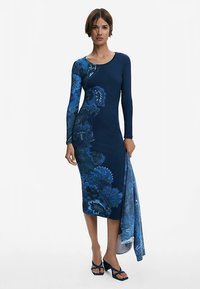 Niebieska sukienka z długim rękawem w kwiatowe wzory, dopasowany krój, długość do kolan. Dopełniona pasującym szalem, zestawiona z niebieskimi sandałami na wysokim obcasie.