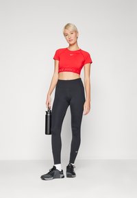Top roșu scurt cu un logo, colanți negri cu talie înaltă, pantofi sport negri și o sticlă de apă neagră în mână.