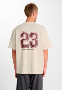 Cremefarvet oversized t-shirt med stort, bordeauxrødt "23" og teksten "INTERNATIONALS" trykt på ryggen. Korte ærmer, afslappet pasform.
