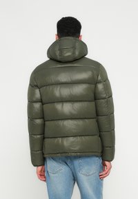 Moški obrnjen stran wearing olive green puffer jacket s kapuco in svetlo modre kavbojke ob navadni beli ozadju.