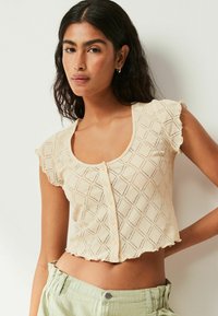 Haut court à manches courtes en beige clair, avec un motif de diamants, un col rond et une fermeture à boutons, aux bords volants.
