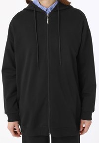 Black zip-up hoodie with a spacious fit, hood, drawstrings, and ribbed cuffs. Smooth fabric texture and a straight hemline.