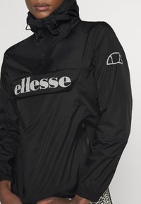 Osoba nosząca czarną kurtkę przeciwwiatrową z logo "ellesse" na klapie przedniej kieszeni i małym logo na lewym rękawie.
