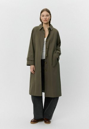 Olivegrüner langer Mantel mit lockerer Passform, ausgestattet mit einem Kragen, Knopfverschluss und Seitentaschen. Kombiniert mit dunkelblauen weit geschnittenen Jeans.