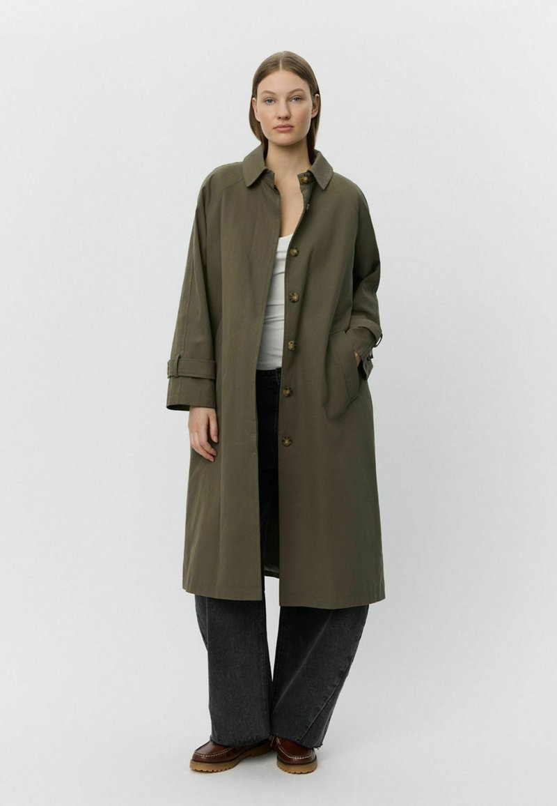 Olíva zöld, laza szabású, hosszú kabát, galléros nyakkal, gombos záródással és oldalsó zsebekkel. Sötét, bő szárú farmernadrággal párosítva.