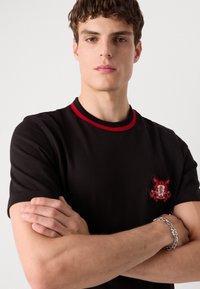 Black short-sleeve t-shirt with a red ribbed collar, featuring a textured emblem on the chest and a silver chain bracelet on the wrist.