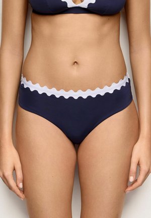 Kobieta nosząca granatowe majtki od bikini z białą falistą lamówką, pokazana od środka tułowia do górnej części ud na jednolitym tle.