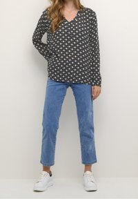 Kobieta ubrana w czarną i białą koszulę z geometrycznym wzorem i długim rękawem, niebieskie dżinsy i białe sneakersy, stojąca z jedną ręką na biodrze.