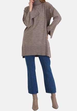 Femme portant un pull à col roulé oversize taupe, un jean court bleu et des bottines taupe, posant avec la main près du visage et de la hanche.