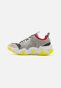 Zapatillas deportivas en tonos gris y blanco con una suela gruesa y detalles amarillos, paneles de malla y un collar acolchado en gris y rojo. Nombre de la marca visible.