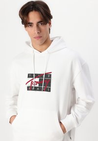 Biely mikina s prednou kengurou vreckom, ktorá na hrudi obsahuje čierno-červený grafický logo "Tommy Jeans" a šnúrky na kapucni.