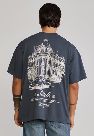 Férfi, aki sötétkék pólót visel, amelynek hátoldalán egy vintage épület, autó, pálmafa és dőlt betűs "SikSilk" felirat látható.