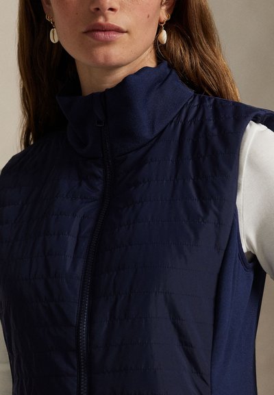 Μπλε σκούρο αμάνικο γιλέκο με ψηλό γιακά, με καπιτονέ υφή και φερμουάρ στο μπροστινό μέρος. Φοριέται πάνω από λευκό μακρυμάνικο τοπ.