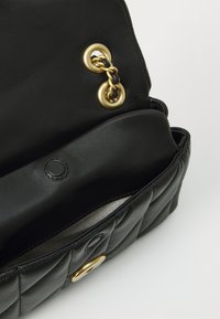 Sac à main en cuir noir avec un design matelassé, quincaillerie dorée, accents en chaîne et un compartiment ouvert révélant un intérieur lisse et un bouton circulaire.