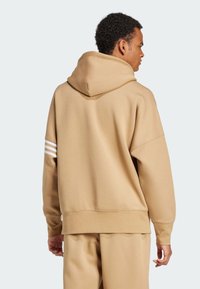 Sudadera con capucha en color marrón claro, de corte relajado, que presenta tres franjas blancas en la manga izquierda y una textura suave. Puños acanalados visibles.