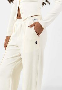 Crème kleurige geribbelde broek met een elastische tailleband, zijzakken en een klein zwart logo op de zak. De stof lijkt zacht en rekbaar.