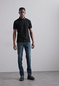 Svart polotröja, blå jeans och svarta skor. Slim fit, kortärmad och med krage med knappdetalj. Enkla ytor, inga mönster.