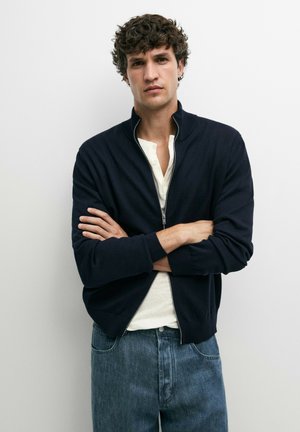 Junger Mann mit lockigem Haar, der einen schwarzen Pullover mit Reißverschluss über einem weißen Hemd und blauen Jeans trägt und mit verschränkten Armen vor einer weißen Wand steht.