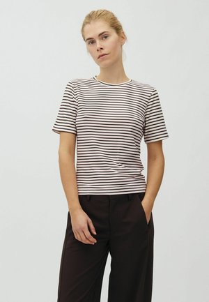 Femme aux cheveux blonds portant un t-shirt à manches courtes rayé et un pantalon noir, debout avec une main dans la poche sur un fond uni.
