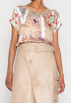 Nainen, joka pukeutunut beigen väriseen, lyhythihaiseen kukkakuosiseen paitaan, jossa on lintuja ja palmuja, ja joka on pantu beigen väriseen, vyölliseen nahkahameeseen, jossa on napit ja tasku.