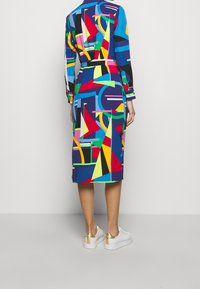 Färggdress med geometriskt mönster i olika former, med en medellång skärning och långa ärmar, tillverkad av mjukt tyg. Bärs med vita skor.