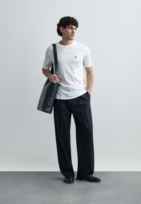 Vyras su garbanotais plaukais, dėvintis baltą marškinėlį ir juodus plačių kojų kelnes, ant peties nešiojantis didelę juodą rankinę, stovintis ant pilkos grindų dangos.