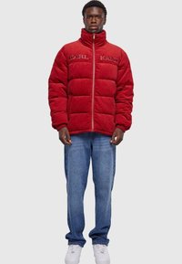 Rød fløjl pufferjakke med høj krave og broderet "KARL KANI" logo, parret med blå jeans og hvide sneakers.
