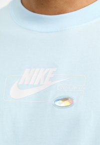 Světle modré bavlněné tričko s bílým potištěným logem Nike a sloganem, s holografickým oválným detailem u lemu.