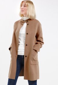 Brauner Wollmantel mit rundem Kragen, Knopfverschluss, zwei Taschen und lockerer Passform. Kombiniert mit einem weißen Rollkragenpullover und blauen Jeans.