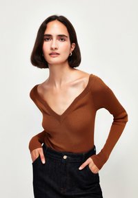 Hnědý žebrovaný bodysuit s dlouhým rukávem a hlubokým výstřihem do V, v kombinaci s tmavými džínovými kalhotami s vysokým pasem.