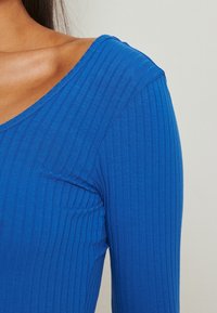 Femme portant un haut bleu vif à manches longues côtelé avec un large décolleté, laissant apparaître une partie de son épaule et de sa clavicule.