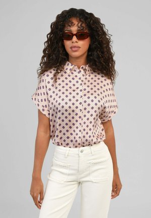 Femme aux cheveux bouclés portant des lunettes de soleil marron, un chemisier à manches courtes à motifs et un pantalon blanc taille haute sur un fond gris clair.