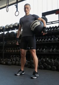 Atletický muž v přiléhavém šedém tričku s krátkými rukávy a černých šortkách drží černo-bílý medicinbal o hmotnosti 6 kg, v pozadí jsou kettlebelly.