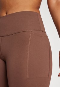 Leggings marron taille haute en tissu lisse et extensible, avec un détail de poche et des coutures contrastantes le long des coutures.