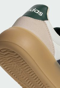 Bílá teniska se zelenými koženými doplňky, krémovou semišovou částí a béžovou gumovou podrážkou s texturovaným vzorem běhounu. Na patě je viditelné logo.