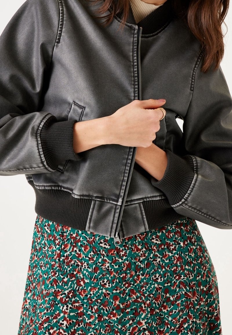 Femme portant une veste bomber gris foncé avec des poignets et un ourlet côtelés, sur une robe ou une jupe à motif floral multicolore.