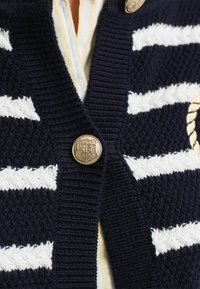 Marinblå cardigan med vita randiga mönster, med en texturerad stickning och en rund guldfärgad knapp med graverade logodetaljer.