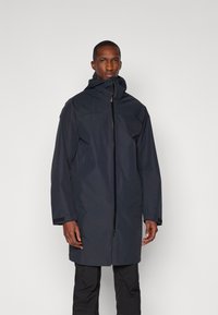 Veste pour homme bleu marine foncé, en matériau imperméable, design avec capuche, fermeture éclair complète, manches longues avec poignets réglables, style minimaliste, sans logos visibles.